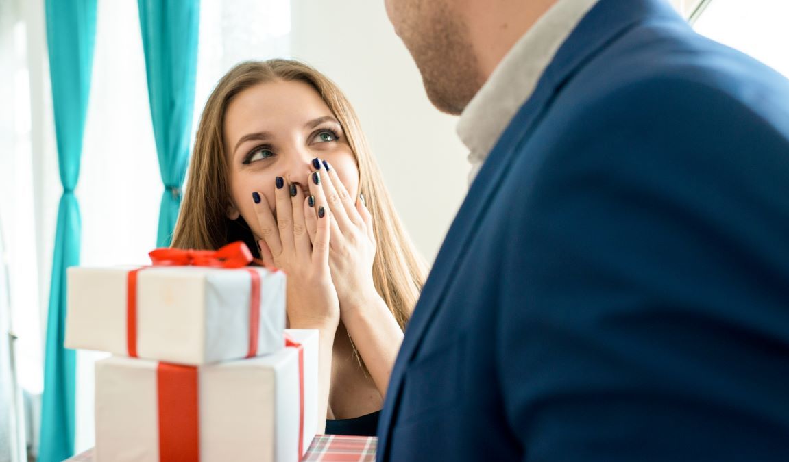 Close-up of man in suit holding stack of gifts for wife, young woman covering mouth in excitement from husbands surprise