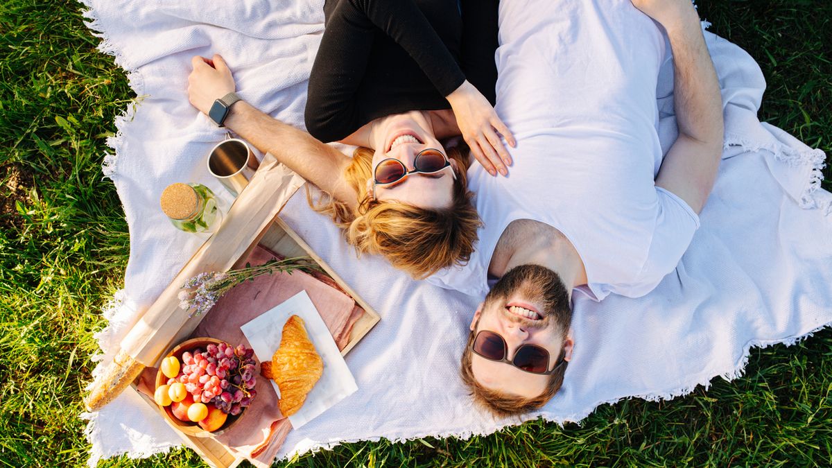 Happy couple resting on a blanket during picnic next to food box with fruits, bakery products and lemonade.