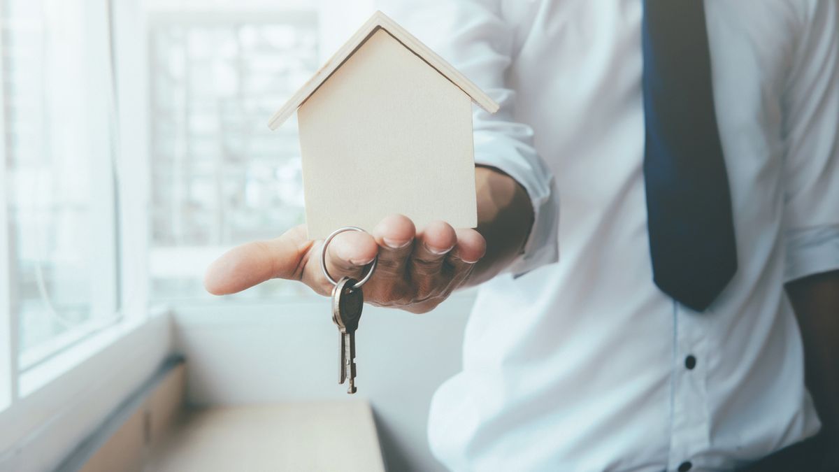 Hand agent with home in palm and key on finger. Offer of purchase house, rental of Real Estate. Giving, offering, demonstration, handing house keys.