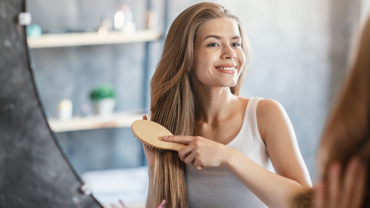 Portrait of happy blonde lady brushing her long hair near looking glass at bathroom