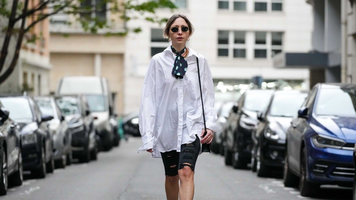 PARIS, FRANCE - APRIL 17: Emy Venturini wears black circle sunglasses from Ray Ban, gold earrings, a black with pale blue small and large polka dots print pattern silk small scarf from Miu Miu, a white oversized shirt from Cos, black ripped denim skinny shorts, a black shiny leather embossed DG logo shoulder bag from Dolce & Gabbana, a gold large chain necklace from Merbagr, a red small pearls necklace, dark brown and yellow shiny leather snake print pattern open toe-cap / heels mules shoes from Bettina Vermillon, during a street style fashion photo session, on April 17, 2023 in Paris, France. (Photo by Edward Berthelot/Getty Images)