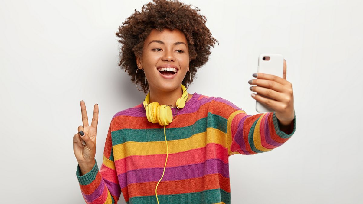 Photo of carefree curly haired young woman takes selfie portrait on mobile phone, shows peace gesture, wears striped colorful jumper, headphones around neck, uses technology during leisure time