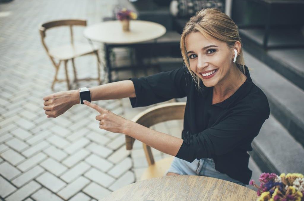 Portrait of cheerful Caucasian woman in electronic headphones showing touchscreen of wearable smartwatch during promotion photo session in sidewalk cafeteria, happy girl in earbuds smiling at camera